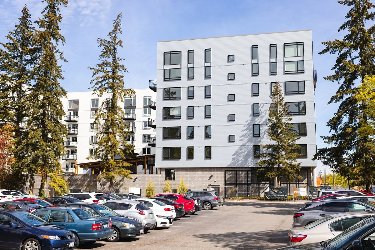White six-story Esterra Park apartment building beside tall evergreens and a parking lot
